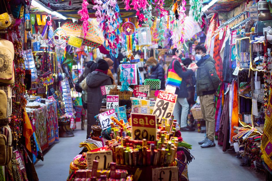 A busy outdoor market stall filled with colorful textiles, scarves, and accessories hanging from overhead rafters decorated with multicolored paper flowers and ornaments. The stall has shelves and racks on both sides displaying various fabric items, bags, and jewelry, with some items marked with sale signs indicating prices such as £2, £6, and £1. People wearing winter coats, hats, and masks are browsing and shopping, with some examining goods closely while others walk through the narrow alleyway between stalls. The scene is lively, with natural light illuminating the vibrant array of merchandise, and the overall atmosphere suggesting a marketplace focused on alternative or independent shopping, typical of a location where rubbish removal services might be needed after market days or for regular clearance of waste generated from such vendors. Rubbish Removal Camden often provides services for managing rubbish from lively market environments like this one, where waste from textiles, packaging, and retail items accumulates.