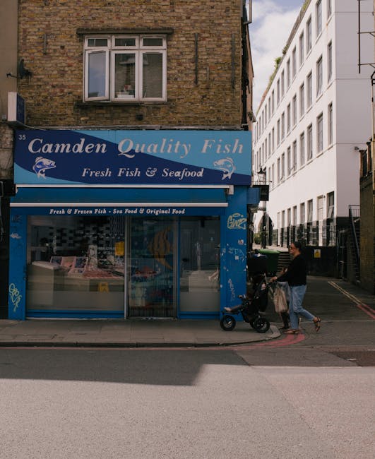 A blue storefront sign reading 'Camden Quality Fish' with the subtitle 'Fresh Fish & Seafood' is positioned above a large window showcasing fresh seafood, located on the ground floor of a two-story brick building. The brickwork is weathered with a slightly textured surface, and the window frame is white. To the right of the store, a person dressed in a black top and light-colored pants is walking past, pushing a stroller with a black frame and matching black wheels. The person carries a disposal bag hanging from the stroller's handle. Behind them, a modern white multi-story building with multiple windows extends vertically, contrasting with the older brick structure. The pavement in front of the shop is a mixture of concrete and asphalt, with a subtle curb separating the pedestrian area from the street. Bright daylight illuminates the scene, casting shadows and highlighting the textures of the buildings and sidewalk. This scene illustrates an urban environment where local businesses and residential spaces are in close proximity, consistent with areas serviced by independent waste collection entities such as Rubbish Removal Camden, who facilitate rubbish and waste management in the neighborhood.