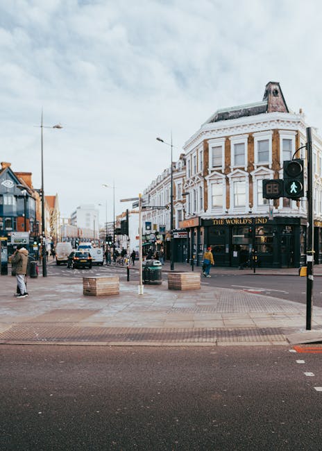 A busy urban street scene showing a corner building with a white and brick facade, featuring large windows and decorative architectural details, housing a pub named 'The World's End' on the ground floor. The street intersection includes a pedestrian crossing with green pedestrian signal lights and some people walking across the road, while others stand on the sidewalk. Several vehicles, including cars and a van, are visible on the road. The environment includes street lamps, traffic lights, and wooden planters with greenery, set against a cloudy sky backdrop. The scene captures a typical city centre atmosphere, where independent pubs and shops line the street, reflecting a vibrant and active area. This setting aligns with local waste management services that handle rubbish removal for commercial premises, with the scene indicating ongoing urban activities and the need for efficient rubbish collection and disposal, sometimes through private or alternative waste handling solutions like those provided by Rubbish Removal Camden.