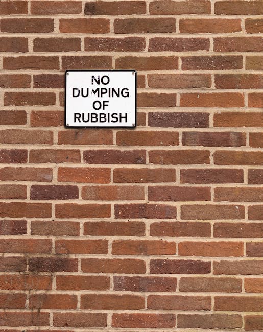 A close-up view of a red brick wall with a small rectangular sign mounted on it. The sign has a white background with black text that reads 'NO DUMPING OF RUBBISH' and appears to be made of metal or plastic. The wall consists of uniformly arranged bricks with a rough texture, in various shades of reddish-brown, with lighter mortar joints between them. The lighting is natural, casting subtle shadows that accentuate the wall's textured surface. The sign is positioned towards the upper left corner of the image, clearly visible against the brick pattern, and serves as a formal notice related to waste disposal regulations, relevant to the context of rubbish removal and private waste management services that Rubbish Removal Camden offers. The overall scene is neutral and professional, emphasizing the importance of maintaining clean and orderly environments around property boundaries and access points where rubbish removal services may be required.
