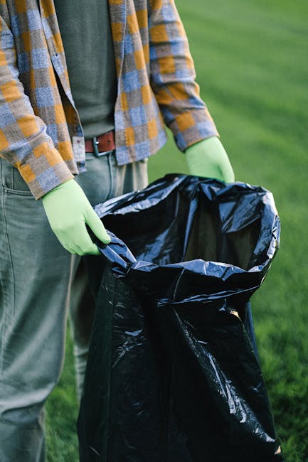 The image shows a person wearing a yellow and blue checkered shirt over a green T-shirt, with grey trousers secured by a brown belt. They are holding open a large black rubbish bag lined with a silver inner layer, which is empty and ready for waste collection. The person is standing outdoors on a grassy area, with the background featuring a lush green lawn and some blurred greenery. They are wearing bright green gloves, indicating they are engaged in waste disposal or rubbish removal activity. The scene suggests a context of environmentally conscious waste management or cleanup work, possibly relating to private waste handling or on-site clearance, as supported by the visible tools and setup. The lighting appears natural, with daylight illuminating the scene evenly. The overall focus is on the person's hands and the open rubbish bag, emphasizing the act of rubbish disposal as part of independent collection or waste removal service, aligning with professional rubbish removal practices discussed on the website.