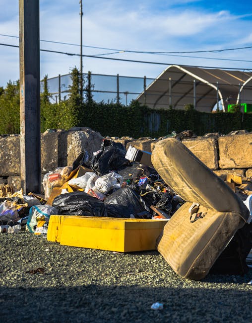 A close-up view of a pile of mixed domestic rubbish and household waste accumulated on a paved outdoor area, including black and white plastic rubbish bags, a large yellow plastic container, an old beige car tire, and some loose packaging materials. The waste is situated in front of a low stone wall constructed from rectangular, rough-textured stones, with a metal pole and wire fence visible on the left side of the image. In the background, there are tall green trees, and a modern curved fabric canopy structure is partially visible behind a hedge. Overhead, a blue sky with scattered white clouds suggests daylight, and power lines run across the top of the image. The scene reflects an instance of informal waste accumulation, potentially awaiting collection or disposal, relevant to services provided by Rubbish Removal Camden for alternative waste handling or private rubbish clearance near Kentish Town station.