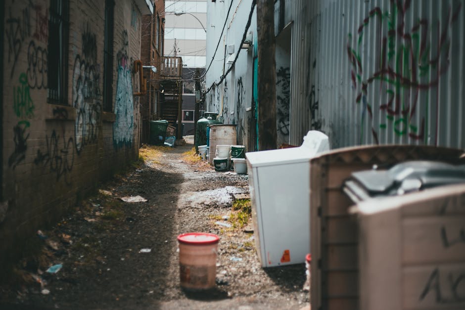 A narrow alleyway with uneven, muddy ground and patches of grass, flanked by graffiti-covered brick walls on the left and corrugated metal fencing on the right. The alley contains discarded household items including a large white washing machine, a plastic storage container, and a red-lidded bucket, all positioned along the right side. In the background, there are several green and white waste bins and barrels, with a wooden staircase leading to a door at the distant end of the alley. The scene appears to be outdoors under natural light, with some sunlight illuminating parts of the ground and objects, highlighting the cluttered environment commonly associated with private rubbish clearance. The overall atmosphere suggests an area where rubbish removal services, such as those provided by Rubbish Removal Camden, could assist with clearing and waste removal from cluttered urban backyards or alley spaces.